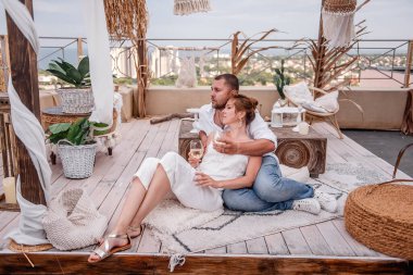 A loving couple sits in a wooden hut in the Moroccan style, drinking white wine. Newlyweds celebrate their anniversary. Young woman laughs, hugs bearded man on open luxury roof terrace with sea view.