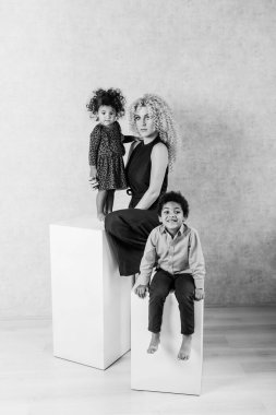 Afrika bukleli beyaz genç anne Afrikalı Amerikalı çocukları kucaklıyor. Stüdyoda gri arka planda beyaz küplerin üzerinde oturan mutlu melez aile. Bir kadın kızını kucağına alıp evlat ediniyor.