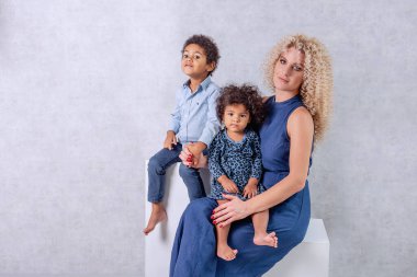 Afrika bukleli beyaz genç anne Afrikalı Amerikalı çocukları kucaklıyor. Stüdyoda gri arka planda beyaz küplerin üzerinde oturan mutlu melez aile. Bir kadın kızını kucağına alıp evlat ediniyor.