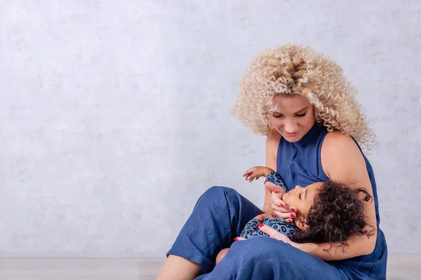 Caucasian young mother with curls hugs African American girl. Happy mixed race family sitting on the wooden floor in the studio on gray background. Woman holds in arms, caress daughter, adoption