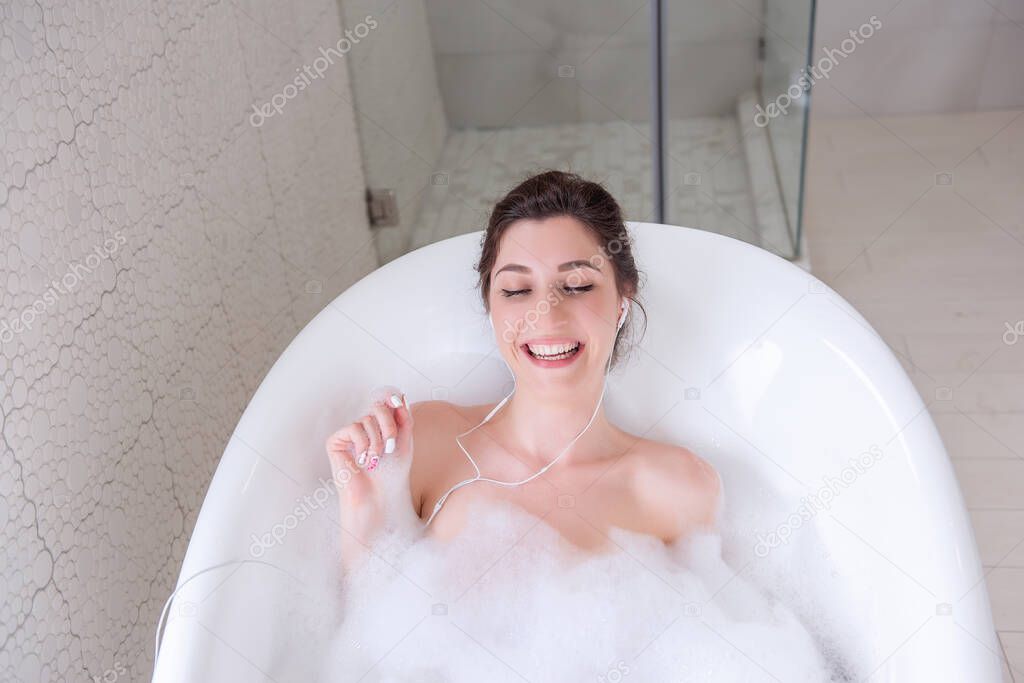 Young millennial woman in headphones listening to music in the bathroom. Happy girl with bun of hair dances in water with white foam, has fun, sings. Spa treatments, relaxing in the morning on weekend