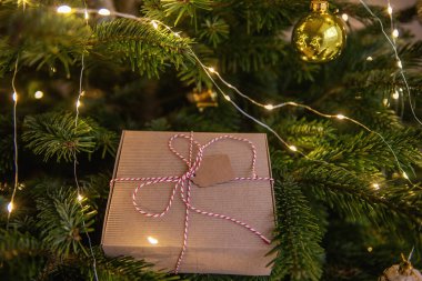 Yeşil bir Noel ağacının dallarında, dilek kartı ile beyaz kırmızı bir sicimle bağlanmış, el sanatları paketinde bir Yeni Yıl hediyesi yatıyor. Parlak çelenk ve oyuncakların şenlikli arka planı. Boşluğu kopyala