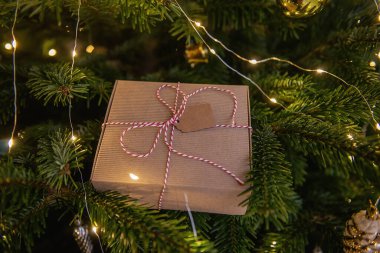 Yeşil bir Noel ağacının dallarında, dilek kartı ile beyaz kırmızı bir sicimle bağlanmış, el sanatları paketinde bir Yeni Yıl hediyesi yatıyor. Parlak çelenk ve oyuncakların şenlikli arka planı. Boşluğu kopyala