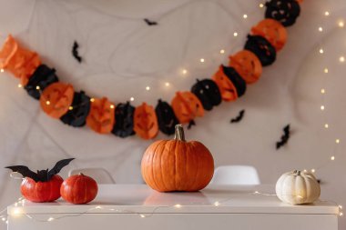 Cadılar Bayramı için zencefilli ve siyah tonlu tebrik kartı. Turuncu balkabakları beyaz bir masada yatar. Örümcek ağları ve siyah yarasaların olduğu kağıt bir çelenk ise arka planda asılı durur. Uzayı kopyala Şenlikli iç mekan