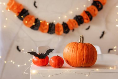 Cadılar Bayramı için zencefilli ve siyah tonlu tebrik kartı. Turuncu balkabakları beyaz bir masada yatar. Örümcek ağları ve siyah yarasaların olduğu kağıt bir çelenk ise arka planda asılı durur. Uzayı kopyala Şenlikli iç mekan