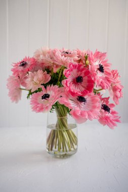 Pembe şakayıklar canlı bir buket olarak düzenlenmiştir. Hassas taç yaprakları ve karanlık merkezler, beyaz bir masada beyaz panelli bir duvara karşı duran cam bir vazoda dururlar.