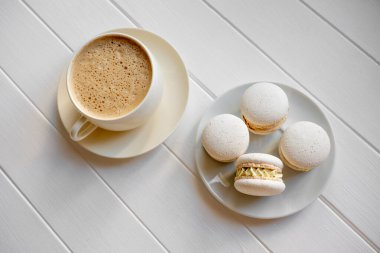 Beyaz bir fincan köpüklü kahvenin yukarıdan görünüşü, küçük bir tabakta dört beyaz badem kurabiyesi, basit, zarif bir kahvaltı veya ahşap bir masada tatlı, minimal ve davetkar.