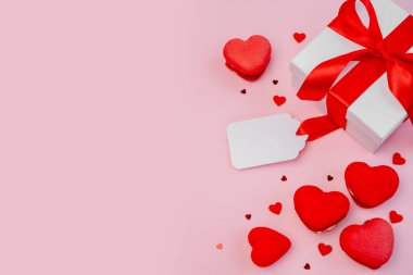 Kırmızı kurdele, kalp şeklinde badem kurabiyesi ve pembe arka planda aşk, romantizm ve özel durumlar için kutlama sembolü olan boş bir etiket ile bağlanmış bir hediye kutusu.