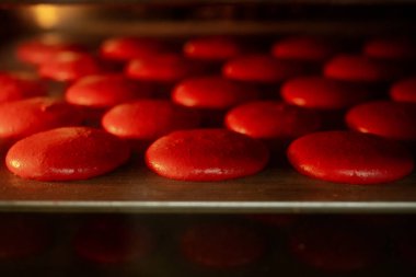 Sıcak fırında metal tepside yükselen kırmızı badem kurabiyeleri. Kabukları kabuklanarak parlak, tatlı Fransız pastalarına dönüşüyor. İçlerinde sıcak altın ışık var.