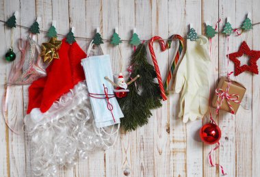 Santa Claus hat and Christmas tree decorations hang on clothespins on a rope against a wooden background with fir branches, Christmas baubles, along with medical masks and gloves.Protect yourself from the coronavirus during Christmas.