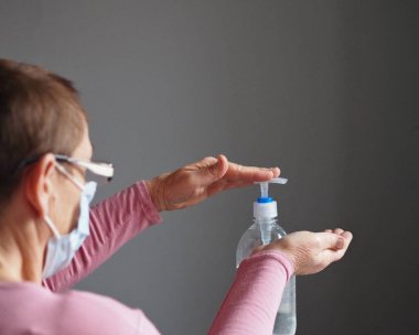 Korunaklı tıbbi maskeli yaşlı bir kadın kendini koronavirüsten korumak için ellerini dezenfekte ediyor. Kişisel antivirüs koruması kullan. Bulanık arkaplan.
