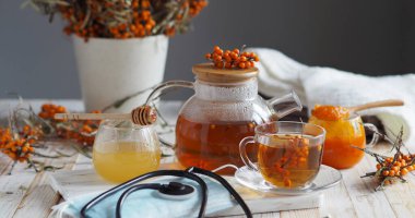 Sağlıklı deniz dikenli böğürtlenleri kullanılarak grip virüsüne karşı koruma ve tedavi kavramı. Koruyucu tıbbi maske, fonendoskop, böğürtlenli deniz dikenli dallar, çay, bal ve beyaz ahşap bir masada sıcak bir eşarp..