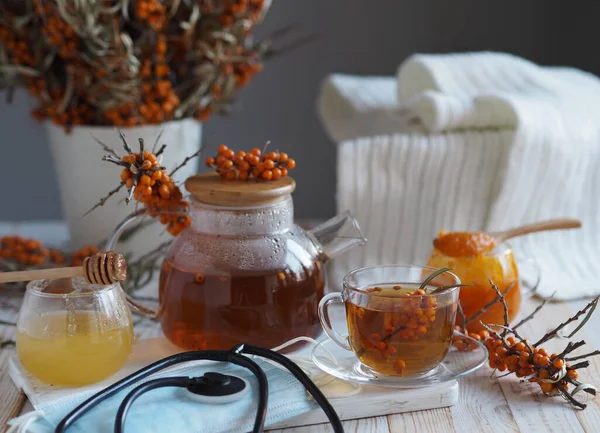 Protective medical mask with a phonendoscope and sea buckthorn branches with berries, tea, honey and a warm scarf on a white wooden table.Concept of protection and treatment of influenza with folk remedies using healthy sea buckthorn berries.