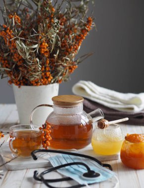 Gribi, sağlıklı deniz karabatağı üzümünü kullanarak tedavi etme fikri. Bir fonendoskoplu tıbbi maske ve böğürtlenli deniz dikenli dallar, çay ve bal ile.