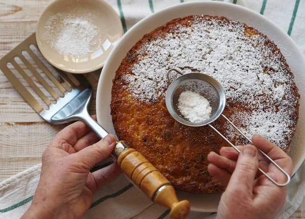 The hands of an elderly woman sprinkle the semolina cake with powdered sugar. The process of making a pie from semolina and dried plums on a wooden background. Food background.
