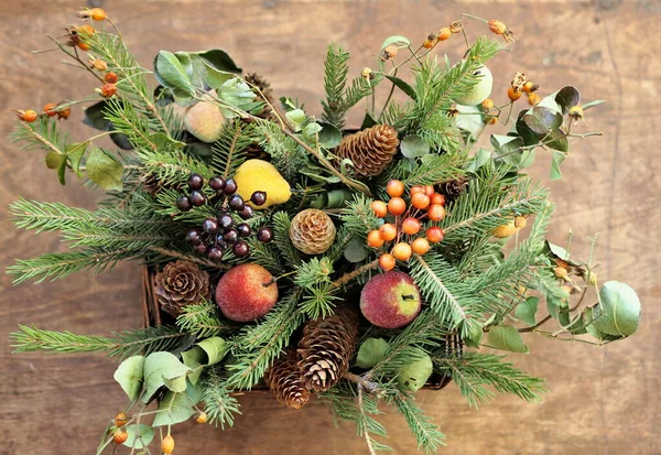 Congratulatory background for cards and congratulations. Bouquet of spruce branches and dry branches of medicinal rose hips with fruit toys in a basket on a wooden background. Nature new year concept.