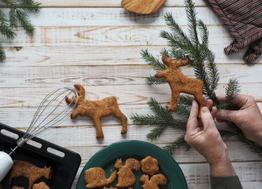 Yaşlı bir kadının elinde tatilde evde pişirilmiş geyik şeklinde bir Noel kurabiyesi var. Bayram için hazırlayıcı çalışmalar. Kurabiye, dekor ve ladin dallarıyla ahşap arka plan..