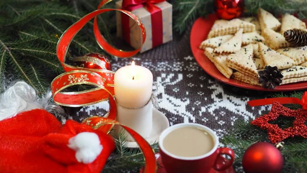 Blurred foreground.Cup of hot coffee with homemade shortbread cookies and red Christmas decor on a knitted knitted background with deer. Congratulatory background. — Stock Photo, Image