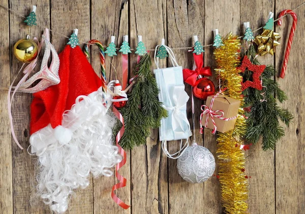クリスマスにコロナウイルスから身を守るんだ サンタクロースの帽子とクリスマスツリーの装飾は 医療用保護マスクと一緒に モミの枝を持つ木製の背景にロープの上に服ピンにかかっています — ストック写真