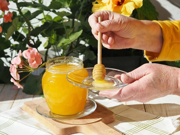 The hands of an elderly woman demonstrate floral natural rustic honey on a background of floral plants. The concept of the health benefits of honey.