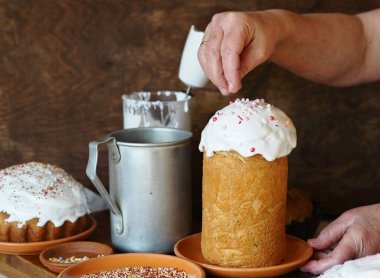 Ev yapımı Paskalya pastası yapma süreci. Bir kadın krem şantili fırınlanmış ürünlerin üzerine pasta süslemesi serpiyor. Köy tarzında ahşap kahverengi arka planda paskalya pastaları..
