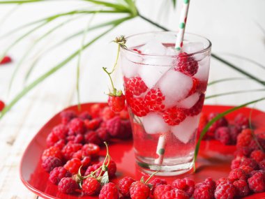 Summer background with a drink. A glass of tonic with raspberries and ice on a red plate. Quench your thirst in hot summer.