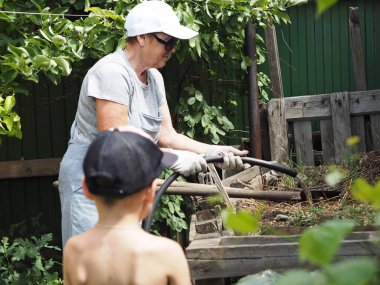 An elderly woman with her grandson is engaged in the preparation of organic fertilizers in private. Naturally recycled homemade compost in a compost heap in the corner of a private garden is hosed with water for the fastest composting.