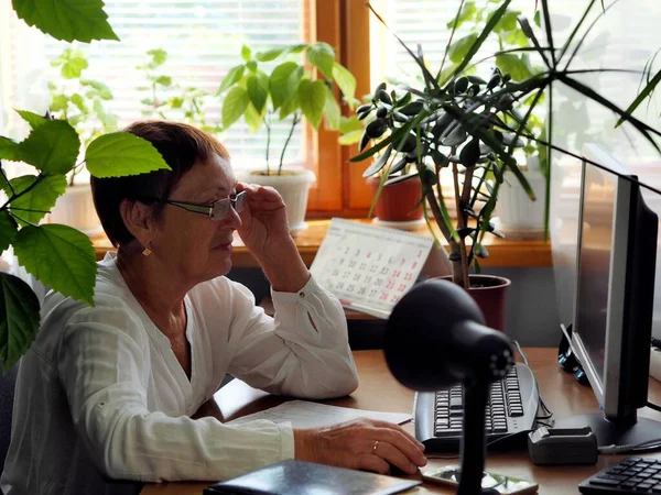 Portrait of an elderly woman correcting her glasses, sitting at a computer, among house plants. Passion for an elderly woman.