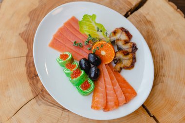 Restoran menüsü. Balık tabağı somon dilimleri, kızarmış yılan balığı, portakal yarımları, marul yaprakları, zeytin ve rulo kırmızı havyarla birlikte doğal ahşap bir arka planda biberiye dalının altında..