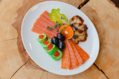 Restoran menüsü. Balık tabağı somon dilimleri, kızarmış yılan balığı, portakal yarımları, marul yaprakları, zeytin ve rulo kırmızı havyarla birlikte doğal ahşap bir arka planda biberiye dalının altında..