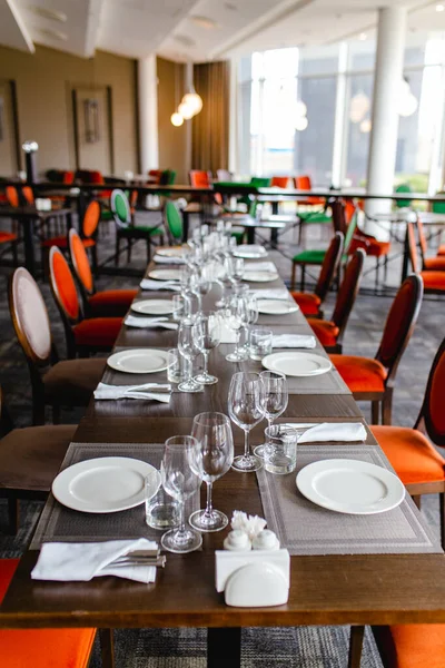 Table setting in a restaurant during a banquet. White empty glass plates, glasses and glasses, cutlery with napkins on the table in a restaurant with green and red upholstered chairs.