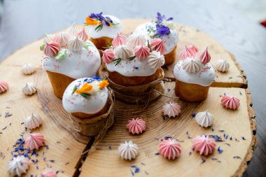 Paskalya dekoru 2021. Beyaz kremalı kekler, pembe kremalı, otlar ve çiçeklerle süslenmiş, ve sepetin içinde doğal ahşap bir arka planda renkli yumurtalar. Paskalya kartı.