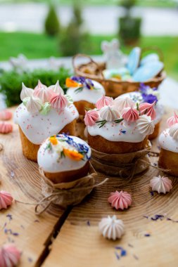 Paskalya dekoru 2021. Beyaz kremalı kekler, pembe bezeli, otlar ve çiçek yapraklarıyla süslenmiş arka planda doğal ahşap arka plan ve yeşillik. Paskalya kartı.