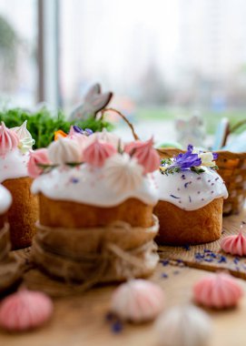 Paskalya dekoru 2021. Beyaz kremalı kekler, pembe bezeli, otlar ve çiçek yapraklarıyla süslenmiş arka planda doğal ahşap arka plan ve yeşillik. Paskalya kartı.