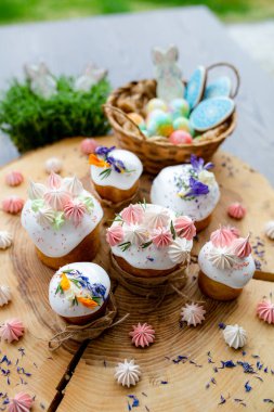 Paskalya dekoru 2021. Beyaz kremalı kekler, pembe bezeli, otlar ve çiçek yapraklarıyla süslenmiş arka planda doğal ahşap arka plan ve yeşillik. Paskalya kartı.