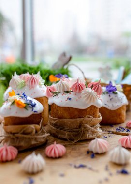 Paskalya dekoru 2021. Beyaz kremalı kekler, pembe bezeli, otlar ve çiçek yapraklarıyla süslenmiş arka planda doğal ahşap arka plan ve yeşillik. Paskalya kartı.