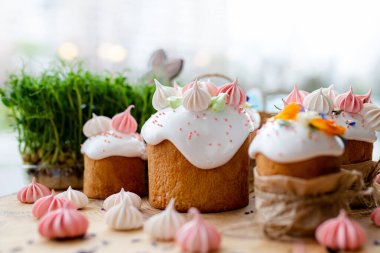 Paskalya dekoru 2021. Beyaz kremalı kekler, pembe bezeli, otlar ve çiçek yapraklarıyla süslenmiş arka planda doğal ahşap arka plan ve yeşillik. Paskalya kartı.