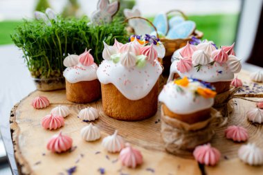 Paskalya dekoru 2021. Beyaz kremalı kekler, pembe bezeli, otlar ve çiçek yapraklarıyla süslenmiş arka planda doğal ahşap arka plan ve yeşillik. Paskalya kartı.