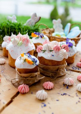 Paskalya dekoru 2021. Beyaz kremalı kekler, pembe bezeli, otlar ve çiçek yapraklarıyla süslenmiş arka planda doğal ahşap arka plan ve yeşillik. Paskalya kartı.