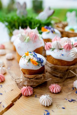 Paskalya dekoru 2021. Beyaz kremalı kekler, pembe bezeli, otlar ve çiçek yapraklarıyla süslenmiş arka planda doğal ahşap arka plan ve yeşillik. Paskalya kartı.