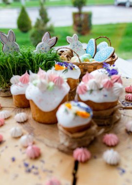 Paskalya dekoru 2021. Beyaz kremalı kekler, pembe bezeli, otlar ve çiçek yapraklarıyla süslenmiş arka planda doğal ahşap arka plan ve yeşillik. Paskalya kartı.