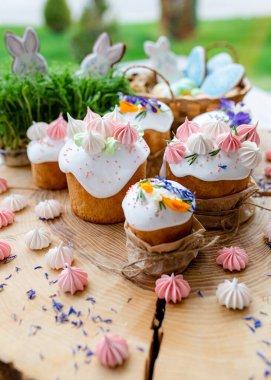 Paskalya dekoru 2021. Beyaz kremalı kekler, pembe bezeli, otlar ve çiçek yapraklarıyla süslenmiş arka planda doğal ahşap arka plan ve yeşillik. Paskalya kartı.