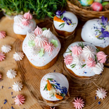 Paskalya dekoru 2021. Beyaz kremalı kekler, pembe bezeli, otlar ve çiçek yapraklarıyla süslenmiş arka planda doğal ahşap arka plan ve yeşillik. Paskalya kartı.