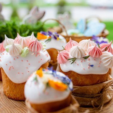 Paskalya dekoru 2021. Beyaz kremalı kekler, pembe bezeli, otlar ve çiçek yapraklarıyla süslenmiş arka planda doğal ahşap arka plan ve yeşillik. Paskalya kartı.