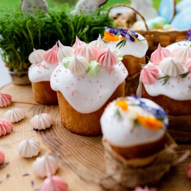 Paskalya dekoru 2021. Beyaz kremalı kekler, pembe bezeli, otlar ve çiçek yapraklarıyla süslenmiş arka planda doğal ahşap arka plan ve yeşillik. Paskalya kartı.