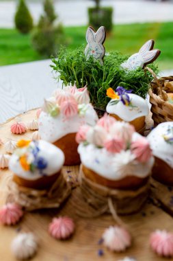 Paskalya dekoru 2021. Beyaz kremalı kekler, pembe bezeli, otlar ve çiçek yapraklarıyla süslenmiş arka planda doğal ahşap arka plan ve yeşillik. Paskalya kartı.