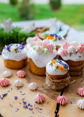 Paskalya dekoru 2021. Beyaz kremalı kekler, pembe bezeli, otlar ve çiçek yapraklarıyla süslenmiş arka planda doğal ahşap arka plan ve yeşillik. Paskalya kartı.