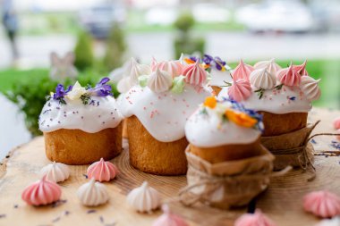 Paskalya dekoru 2021. Beyaz kremalı kekler, pembe bezeli, otlar ve çiçek yapraklarıyla süslenmiş arka planda doğal ahşap arka plan ve yeşillik. Paskalya kartı.