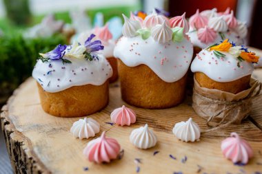 Paskalya dekoru 2021. Beyaz kremalı kekler, pembe bezeli, otlar ve çiçek yapraklarıyla süslenmiş arka planda doğal ahşap arka plan ve yeşillik. Paskalya kartı.