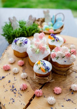 Paskalya dekoru 2021. Beyaz kremalı kekler, pembe bezeli, otlar ve çiçek yapraklarıyla süslenmiş arka planda doğal ahşap arka plan ve yeşillik. Paskalya kartı.